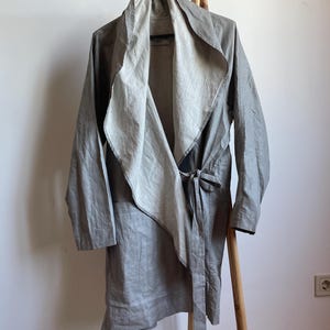 Puede incluir: Una gabardina de lino gris con un diseño largo y fluido. La gabardina tiene un cuello grande que se puede llevar arriba o abajo. La gabardina se ata a la cintura con una faja.