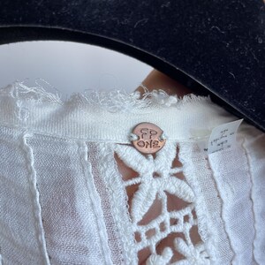 Op de afbeelding: Close-up van een witte blouse met een gedetailleerd bloemen kanten ontwerp. De blouse heeft een kleine, ronde koperen knoop met de tekst "FP ONE". De stof lijkt licht en getextureerd, met een delicaat, open geweven patroon.