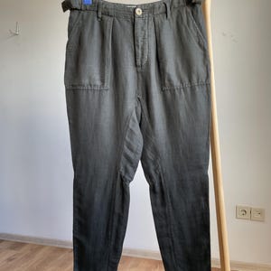 May include: Dark gray pants with a high waist, belt loops, and two front pockets. The pants are hanging on a wooden clothing rack. The fabric appears to have a slight sheen.