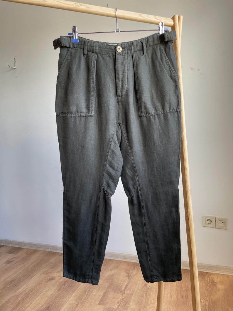 May include: Dark gray pants with a high waist, belt loops, and two front pockets. The pants are hanging on a wooden clothing rack. The fabric appears to have a slight sheen.