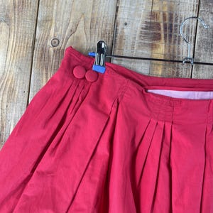 May include: A bright red pleated skirt featuring a high waistband with two matching fabric-covered buttons on the side. The skirt is constructed with vertical pleats that create a full, voluminous silhouette and is suspended from a metal hanger.