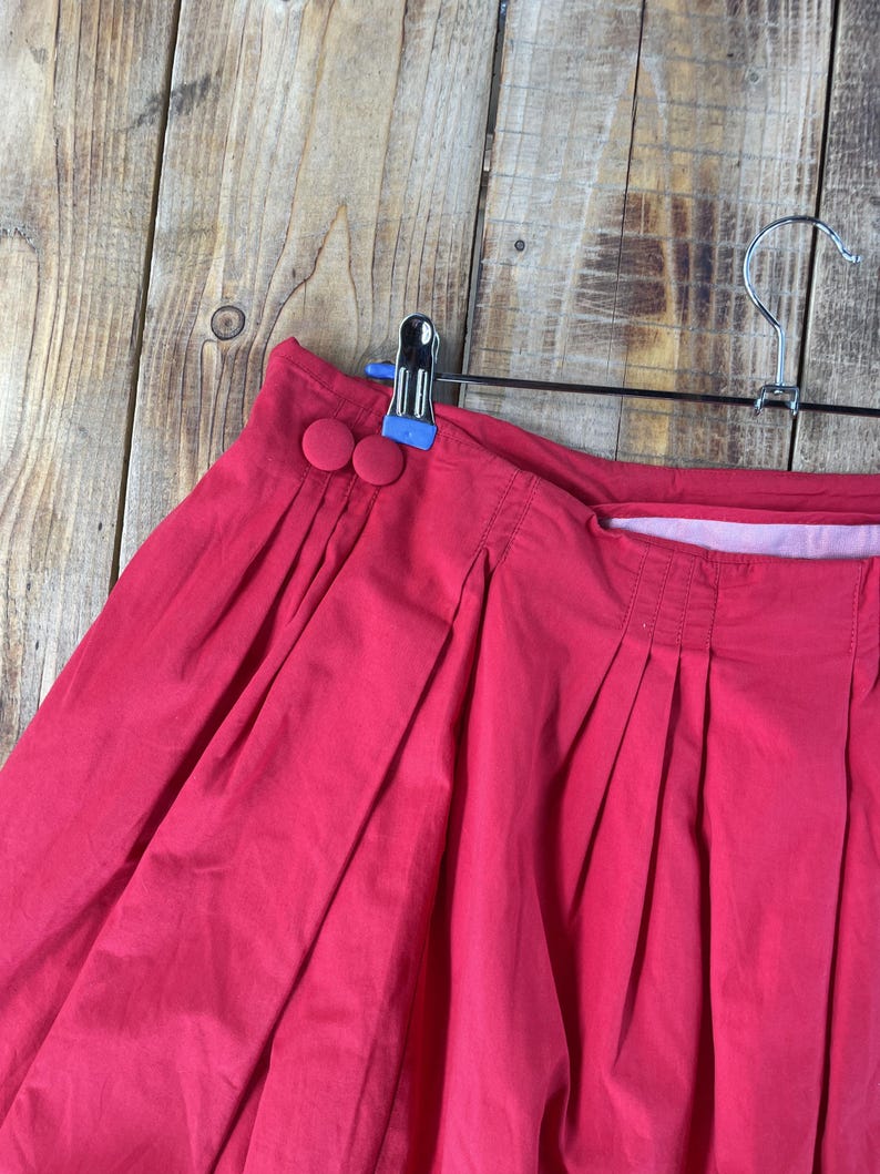 May include: A bright red pleated skirt featuring a high waistband with two matching fabric-covered buttons on the side. The skirt is constructed with vertical pleats that create a full, voluminous silhouette and is suspended from a metal hanger.