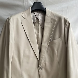 May include: A beige blazer with a notched lapel, a single-button closure, and a chest pocket. The jacket is hanging on a black hanger against a neutral background. The fabric appears to be a lightweight material.