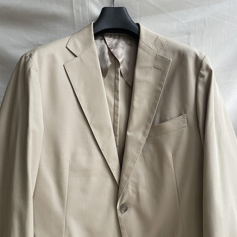May include: A beige blazer with a notched lapel, a single-button closure, and a chest pocket. The jacket is hanging on a black hanger against a neutral background. The fabric appears to be a lightweight material.