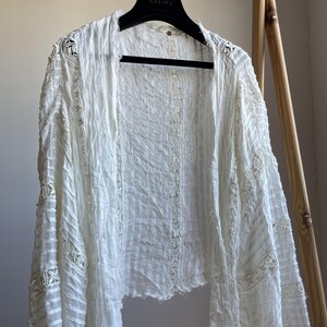 Op de afbeelding: Een witte jas met open voorkant en lange mouwen. De jas heeft een getextureerde, gekreukelde stof en delicate kanten details langs de randen en mouwen. De jas hangt aan een zwarte hanger met de merknaam "CELINE" erop.