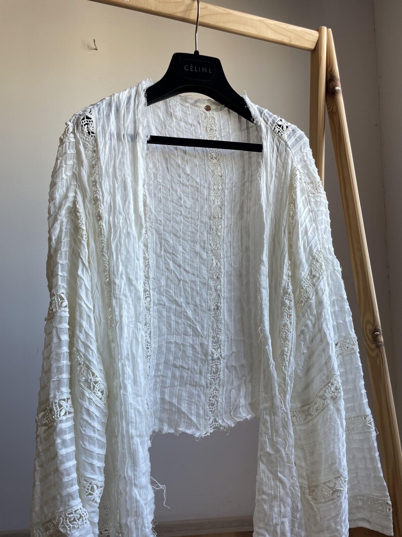 Op de afbeelding: Een witte jas met open voorkant en lange mouwen. De jas heeft een getextureerde, gekreukelde stof en delicate kanten details langs de randen en mouwen. De jas hangt aan een zwarte hanger met de merknaam "CELINE" erop.