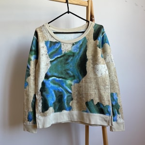 May include: A beige sweatshirt with a blue and green abstract print. The sweatshirt has a round neckline and long sleeves.