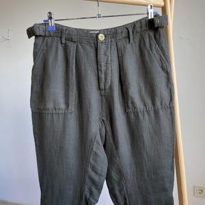 May include: Dark olive green pants with a belt, hanging on a hanger. The pants have two front pockets and a button closure. The fabric appears to be a linen blend. The pants are a neutral color and could be worn with many different tops.