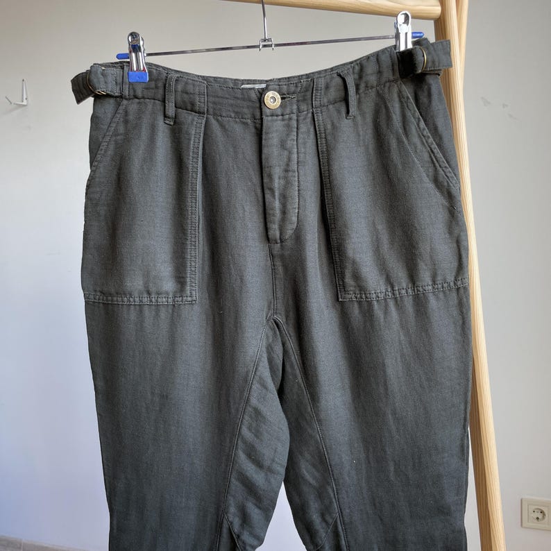May include: Dark olive green pants with a belt, hanging on a hanger. The pants have two front pockets and a button closure. The fabric appears to be a linen blend. The pants are a neutral color and could be worn with many different tops.