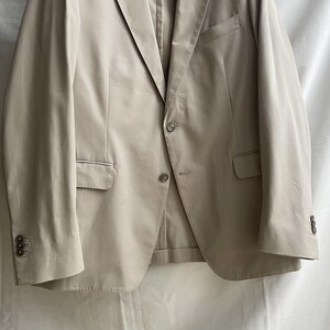 May include: A tan blazer with a two-button closure, notched lapels, and three buttons on each cuff. The blazer has two front pockets and a chest pocket. The fabric appears to be a lightweight material.