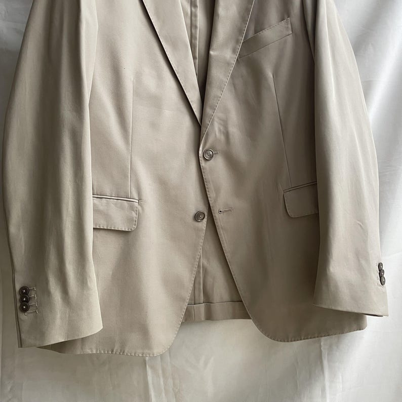 May include: A tan blazer with a two-button closure, notched lapels, and three buttons on each cuff. The blazer has two front pockets and a chest pocket. The fabric appears to be a lightweight material.