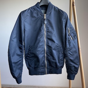 Può includere: Giacca bomber blu navy con cerniera dorata e una piccola tasca con cerniera sulla manica sinistra. La giacca ha colletto, polsini e orlo a coste. È appesa a un attaccapanni di legno.