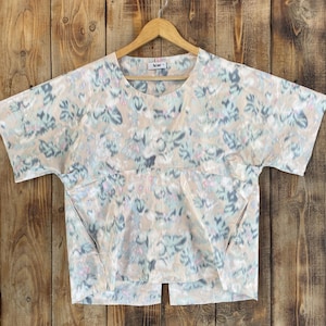 Puede incluir: Una blusa de manga corta con base beige y estampado floral abstracto en tonos azul, verde y rosa. La prenda presenta un cuello redondo, una costura horizontal en el pecho y un dobladillo con abertura. Se observa una etiqueta de Acne Studios en el cuello.