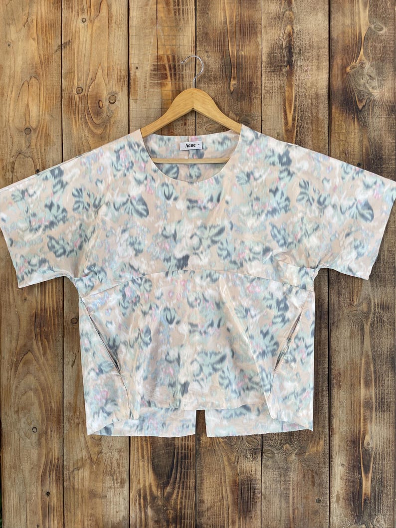 Puede incluir: Una blusa de manga corta con base beige y estampado floral abstracto en tonos azul, verde y rosa. La prenda presenta un cuello redondo, una costura horizontal en el pecho y un dobladillo con abertura. Se observa una etiqueta de Acne Studios en el cuello.