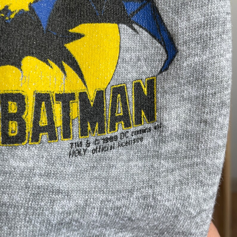 May include: A grey sweatshirt featuring a black and yellow graphic of Batman. The text "BATMAN" is printed in black lettering. The text "TM & &copy; 1989 DC Comics.  JOLAY official licensee" is printed in smaller black lettering below the graphic.
