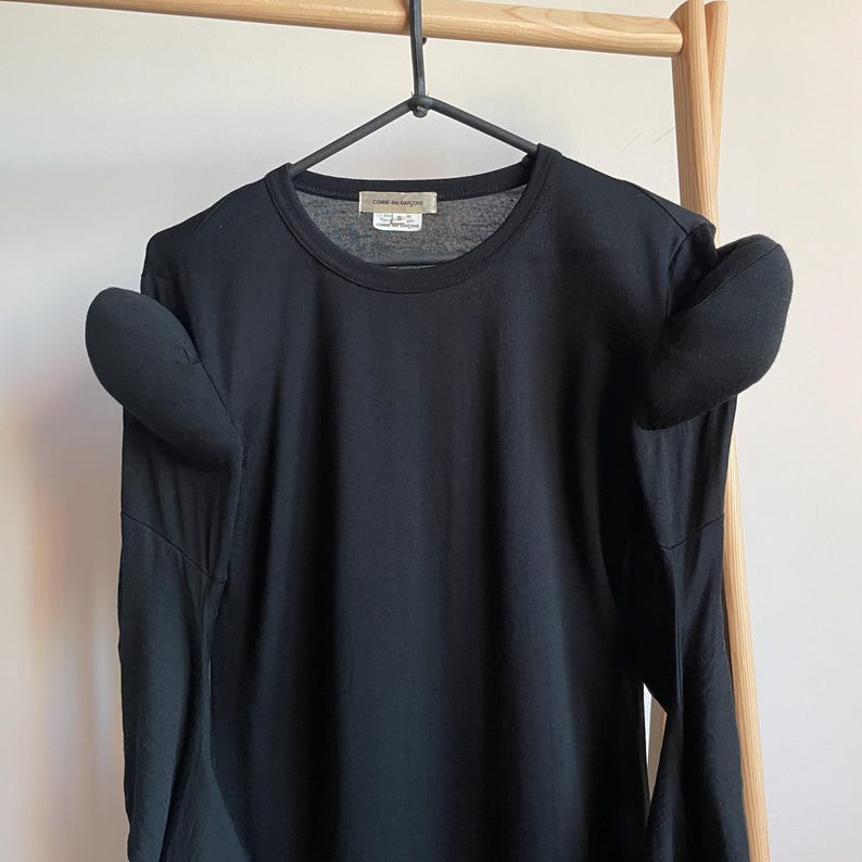 Puede incluir: Camiseta negra de manga larga con cuello redondo y dos alas de tela unidas a los hombros.
