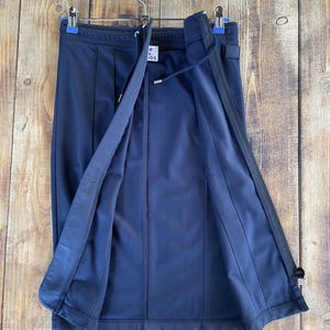 May include: A dark navy blue midi skirt featuring a front zip closure, a drawstring waistband, and vertical seam detailing. The garment includes a white interior label with the text "Acne Studios" and a small square patch on the upper right side.