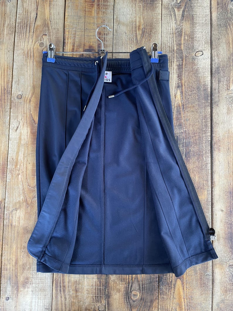 May include: A dark navy blue midi skirt featuring a front zip closure, a drawstring waistband, and vertical seam detailing. The garment includes a white interior label with the text "Acne Studios" and a small square patch on the upper right side.