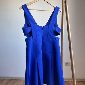 Puede incluir: Un vestido azul vibrante con escote en V y detalles recortados en los laterales. El vestido est&aacute; colgado de una percha de madera, mostrando su dise&ntilde;o y silueta. El vestido tiene cierre de cremallera en la espalda.