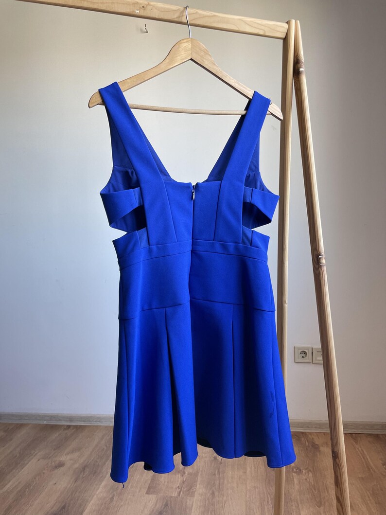 Puede incluir: Un vestido azul vibrante con escote en V y detalles recortados en los laterales. El vestido est&aacute; colgado de una percha de madera, mostrando su dise&ntilde;o y silueta. El vestido tiene cierre de cremallera en la espalda.