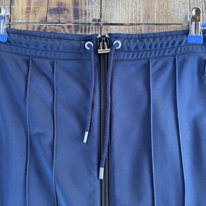 May include: A navy blue pleated skirt featuring a central vertical zip closure and a matching drawstring cord with silver-toned metal aglets at the ends. The garment is suspended from a metal hanger with two blue-tipped clips at the waistband.