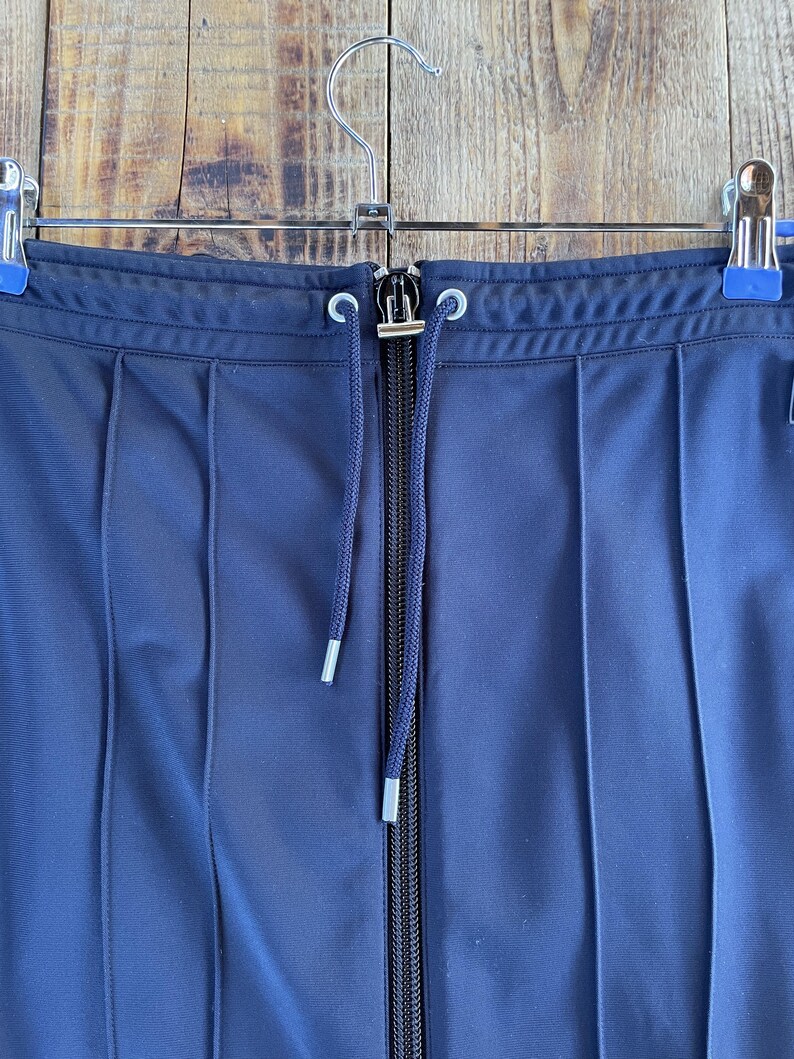 May include: A navy blue pleated skirt featuring a central vertical zip closure and a matching drawstring cord with silver-toned metal aglets at the ends. The garment is suspended from a metal hanger with two blue-tipped clips at the waistband.
