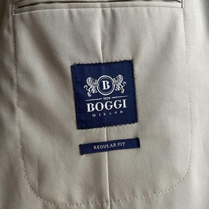 May include: Close-up of the interior of a tan blazer. A navy blue label with the text "BOGGI MILANO 1939" and a "B" logo is stitched inside. Below the label is a smaller navy blue tag that reads "REGULAR FIT".