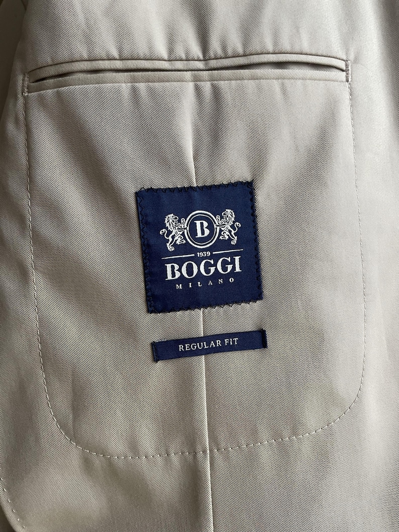 May include: Close-up of the interior of a tan blazer. A navy blue label with the text "BOGGI MILANO 1939" and a "B" logo is stitched inside. Below the label is a smaller navy blue tag that reads "REGULAR FIT".