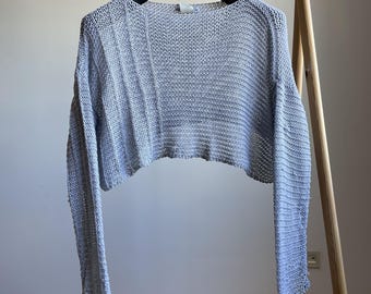 May include: A light gray, open-weave knit sweater with long sleeves. The cropped top hangs from a black hanger on a wooden clothing rack. The sweater has a scoop neck and a slightly textured appearance.
