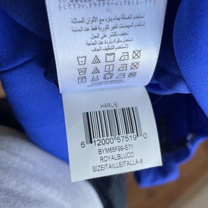 Puede incluir: Etiqueta de prenda azul real con instrucciones de cuidado en varios idiomas. La etiqueta incluye un c&oacute;digo de barras, la palabra "HARLIE" y el c&oacute;digo de producto BYM65F99-E71. La talla se indica como TALLA-8.
