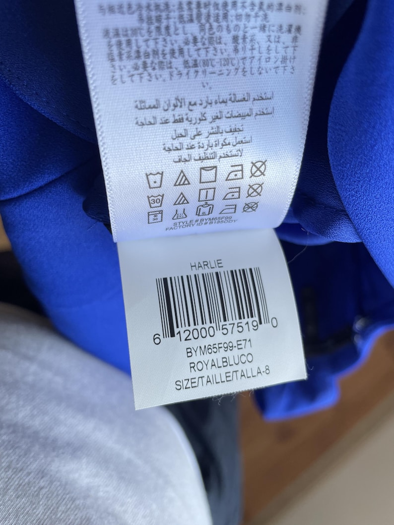 Puede incluir: Etiqueta de prenda azul real con instrucciones de cuidado en varios idiomas. La etiqueta incluye un c&oacute;digo de barras, la palabra "HARLIE" y el c&oacute;digo de producto BYM65F99-E71. La talla se indica como TALLA-8.