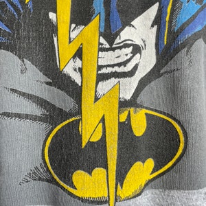 May include: A grey t-shirt featuring a black and yellow Batman logo with a yellow lightning bolt. The Batman logo is enclosed within a black oval.