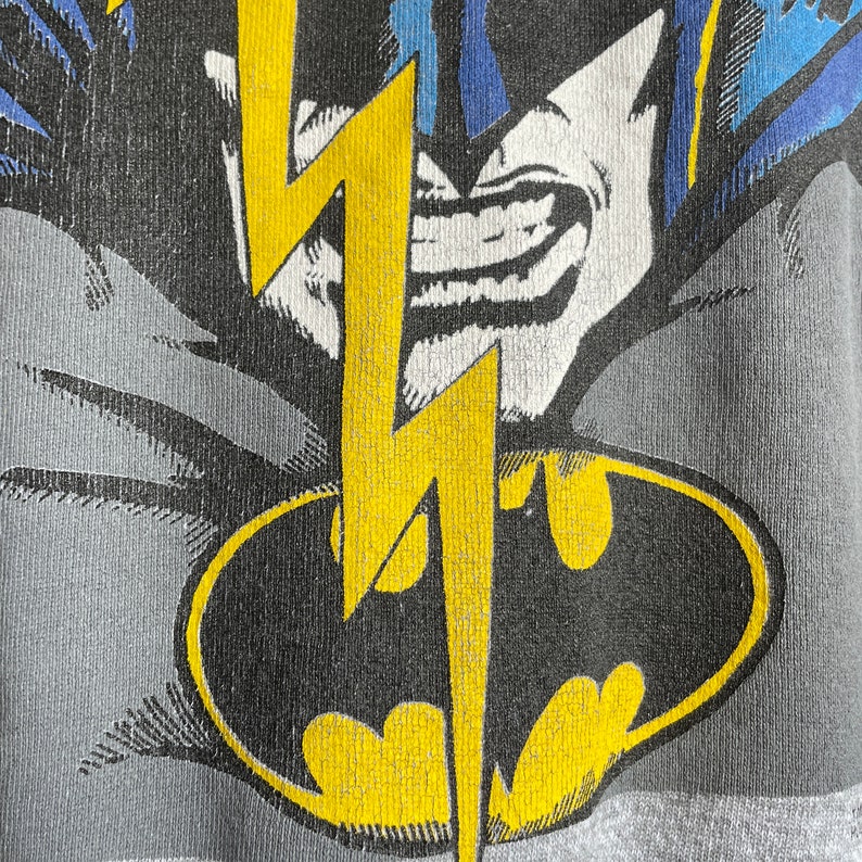 May include: A grey t-shirt featuring a black and yellow Batman logo with a yellow lightning bolt. The Batman logo is enclosed within a black oval.