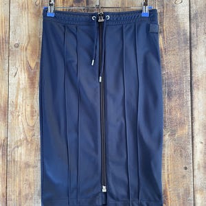 May include: A navy blue knee-length skirt featuring a full-length vertical centre zip, a drawstring waistband with silver-toned metal aglets, and vertical stitched pleat detailing running down the front. A small square patch is on the upper right.