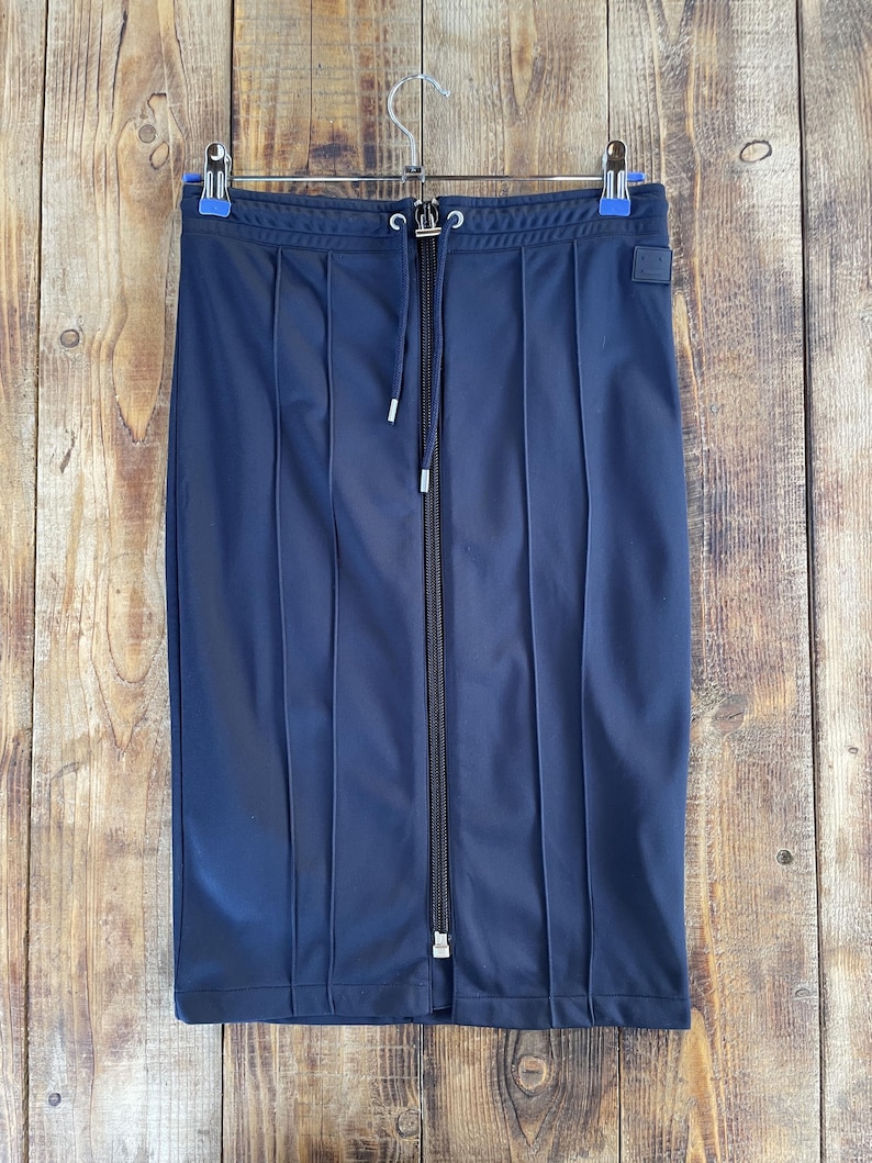 May include: A navy blue knee-length skirt featuring a full-length vertical centre zip, a drawstring waistband with silver-toned metal aglets, and vertical stitched pleat detailing running down the front. A small square patch is on the upper right.
