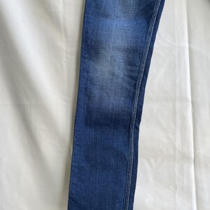 Puede incluir: Primer plano de unos vaqueros de mezclilla azules. Los vaqueros tienen un dise&ntilde;o cl&aacute;sico de cinco bolsillos y un corte recto. La mezclilla tiene un lavado descolorido, con zonas m&aacute;s claras en los muslos y las rodillas. La costura es de color contrastante.