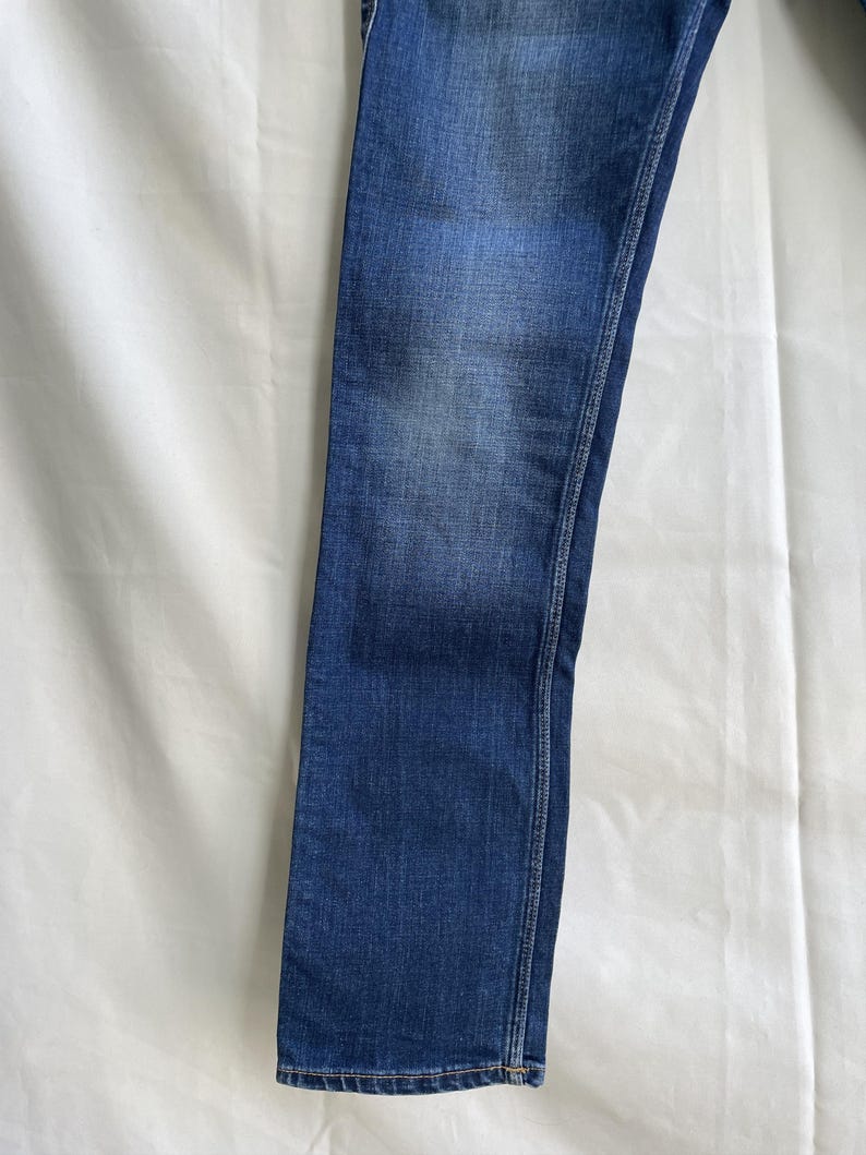 Puede incluir: Primer plano de unos vaqueros de mezclilla azules. Los vaqueros tienen un dise&ntilde;o cl&aacute;sico de cinco bolsillos y un corte recto. La mezclilla tiene un lavado descolorido, con zonas m&aacute;s claras en los muslos y las rodillas. La costura es de color contrastante.