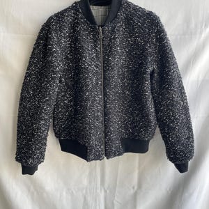 May include: A black and white textured bomber jacket with a full-length zipper. The jacket has black ribbed cuffs and a black ribbed hem. The collar is black with a plaid lining. The jacket is hanging on a black hanger.