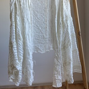 Op de afbeelding: Een witte, openvallende cardigan met lange mouwen, hangend aan een zwarte hanger. De lichtgewicht stof heeft een getextureerde, gekreukelde uitstraling met delicate kanten details langs de mouwen en de voorranden. Het kledingstuk wordt gepresenteerd tegen een neutrale achtergrond.