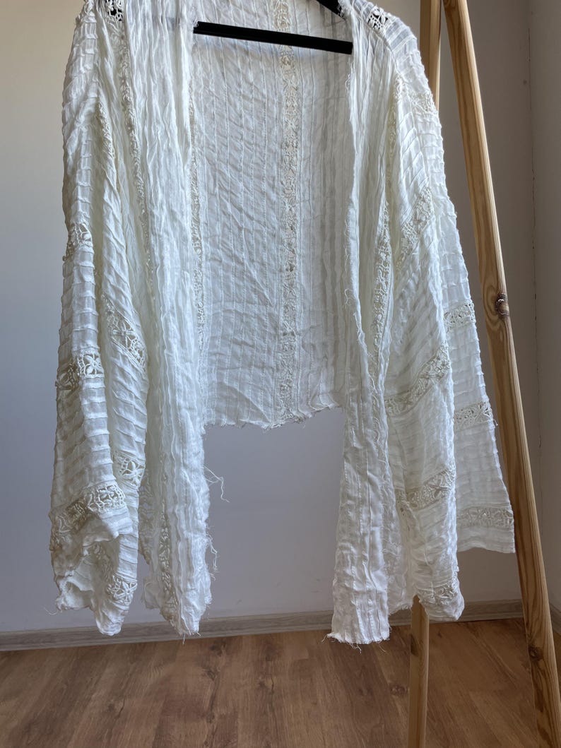 Op de afbeelding: Een witte, openvallende cardigan met lange mouwen, hangend aan een zwarte hanger. De lichtgewicht stof heeft een getextureerde, gekreukelde uitstraling met delicate kanten details langs de mouwen en de voorranden. Het kledingstuk wordt gepresenteerd tegen een neutrale achtergrond.