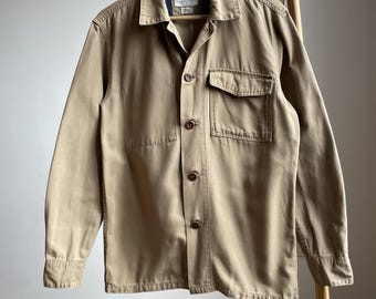 Può includere: Una camicia beige con bottoni, colletto e una tasca sul petto. La camicia è appesa a una gruccia di legno e a un attaccapanni di legno. La camicia ha maniche lunghe e bottoni marroni. Il tessuto è robusto.