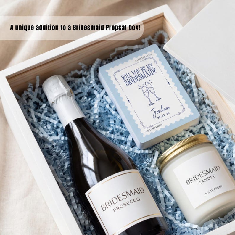 K&ouml;nnte beinhalten: Eine wei&szlig;e Holzgeschenkbox gef&uuml;llt mit hellblauem Papierstreifen, die eine Flasche Bridesmaid Prosecco, eine wei&szlig;e Kerze mit der Aufschrift Bridesmaid Candle White Peony und eine Karte mit der Aufschrift Will You Be My Bridesmaid? und Jordan 09.12.26 enth&auml;lt.