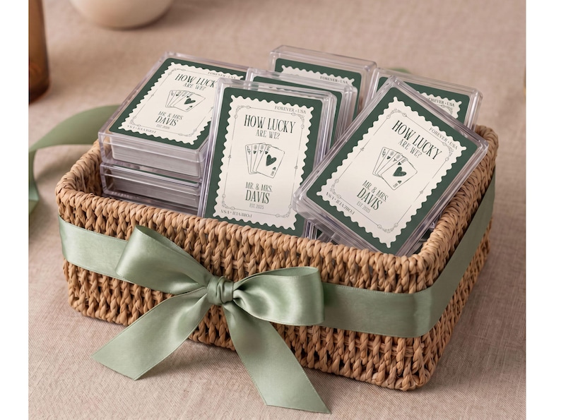 K&ouml;nnte beinhalten: Ein geflochtener Korb mit mehreren durchsichtigen Kunststoffh&uuml;llen f&uuml;r Spielkarten. Jedes Kartenspiel hat einen gr&uuml;nen Rand und den Text "HOW LUCKY ARE WE? MR. & MRS. DAVIS EST. 2023" sowie eine Abbildung von Spielkarten. Ein salbeigr&uuml;nes Band ist um den Korb gebunden.
