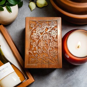 以下が含まれることがあります： 蓋に花柄が彫刻された木製の箱。箱は閉まっており、濃い茶色の仕上げになっています。箱は灰色の表面の上に置かれています。