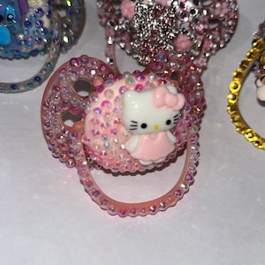 Può includere: Un ciuccio rosa decorato con numerosi piccoli strass iridescenti e una figura centrale di Hello Kitty. Il ciuccio presenta un anello circolare e uno scudo ricoperto di gemme scintillanti, con un piccolo personaggio bianco e rosa applicato.