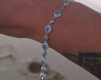 Puede incluir: Una pulsera de plata con una serie de piedras preciosas redondas de color azul claro. Cada gema está engastada en un marco de plata, algunas rodeadas de piedras claras más pequeñas. La pulsera se muestra sobre un fondo borroso.