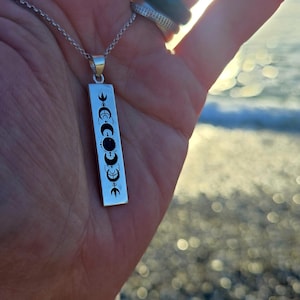Puede incluir: Un collar de plata con un colgante que presenta una barra vertical con un diseño de fase lunar y una piedra preciosa negra en el centro.