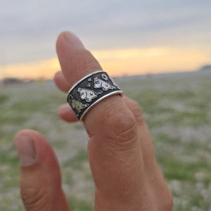 Könnte beinhalten: Silberring mit einem sich wiederholenden Muster von Cartoonfiguren. Der Ring wird an einem Finger getragen, mit einem verschwommenen Hintergrund aus Feld und Himmel. Das Design des Rings umfasst kleine, detaillierte Figuren in einer Kontrastfarbe zum Silberband.