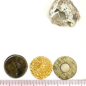 May include: Three round, decorative, flat, disc-shaped objects. The first is a dark brown and gray disc with a white border. The second is a light brown disc with a speckled pattern. The third is a silver disc with a black center and a gold rim. The disc has text in a circular pattern around the edge. A ruler is visible in the bottom of the image.