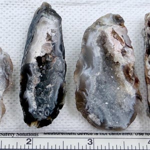 May include: Four polished agate stones with a variety of colors including gray, white, brown, and black. The stones have a natural, organic shape and texture. The image includes a ruler for scale.